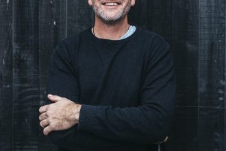 winery owner male standing and smiling with wine bottle next to him
