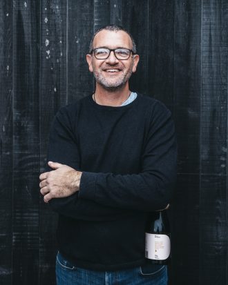 winery owner male standing and smiling with wine bottle next to him