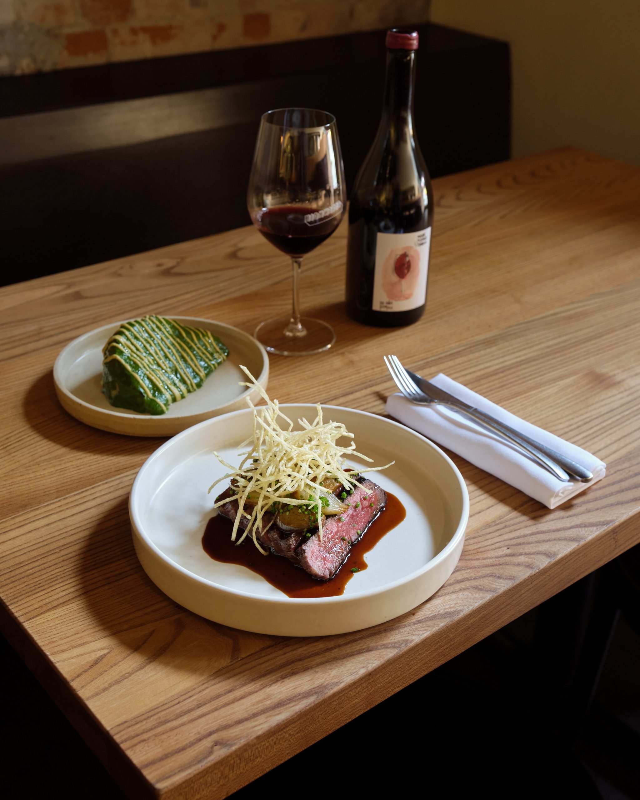 plate of meat and garnish, glass of red wine and bottle, and napkin and cutlery on wooden table