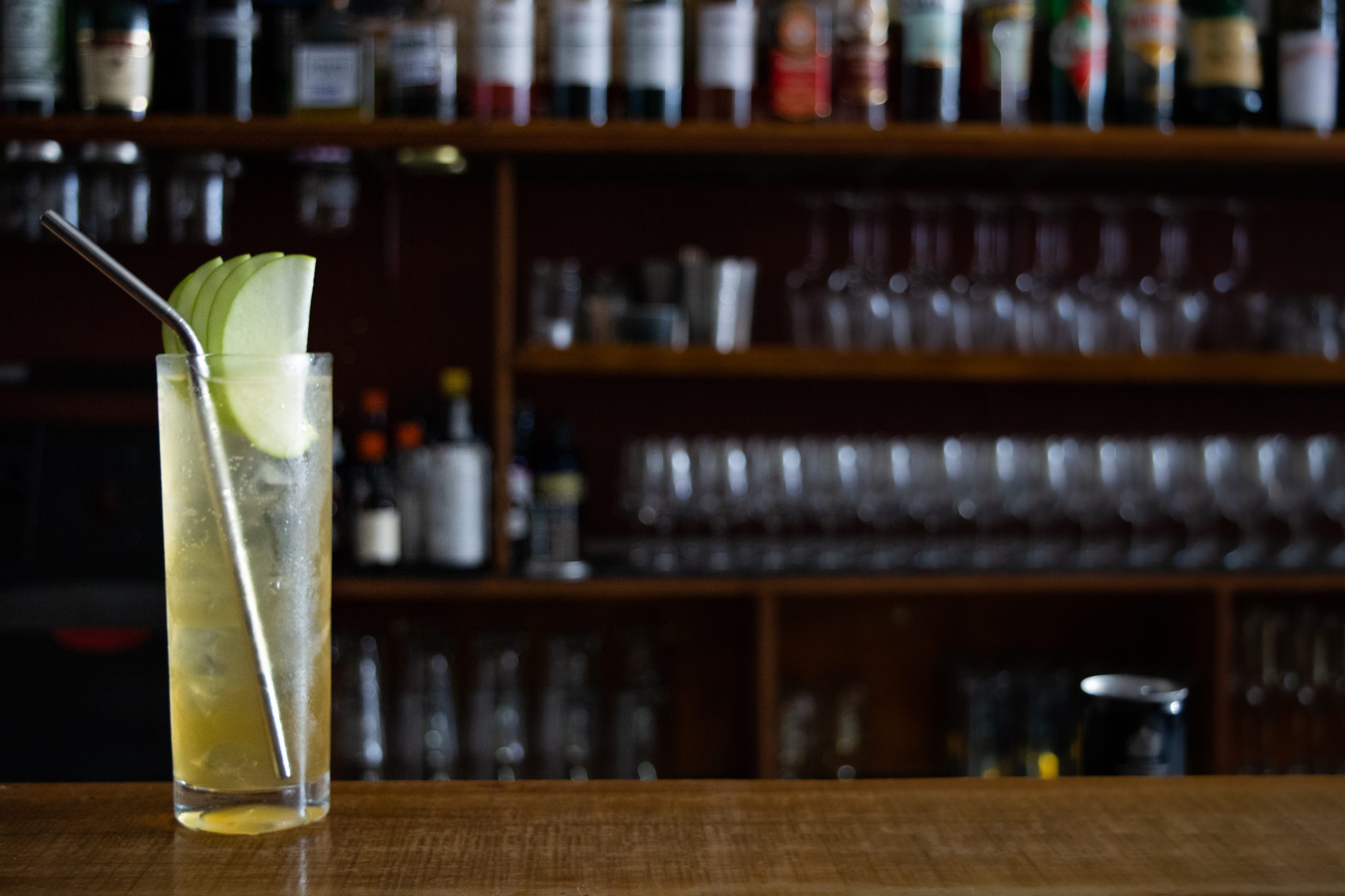 yellow cocktail with lime and metal straw in glass on bar bench with glasses and bottles in the background