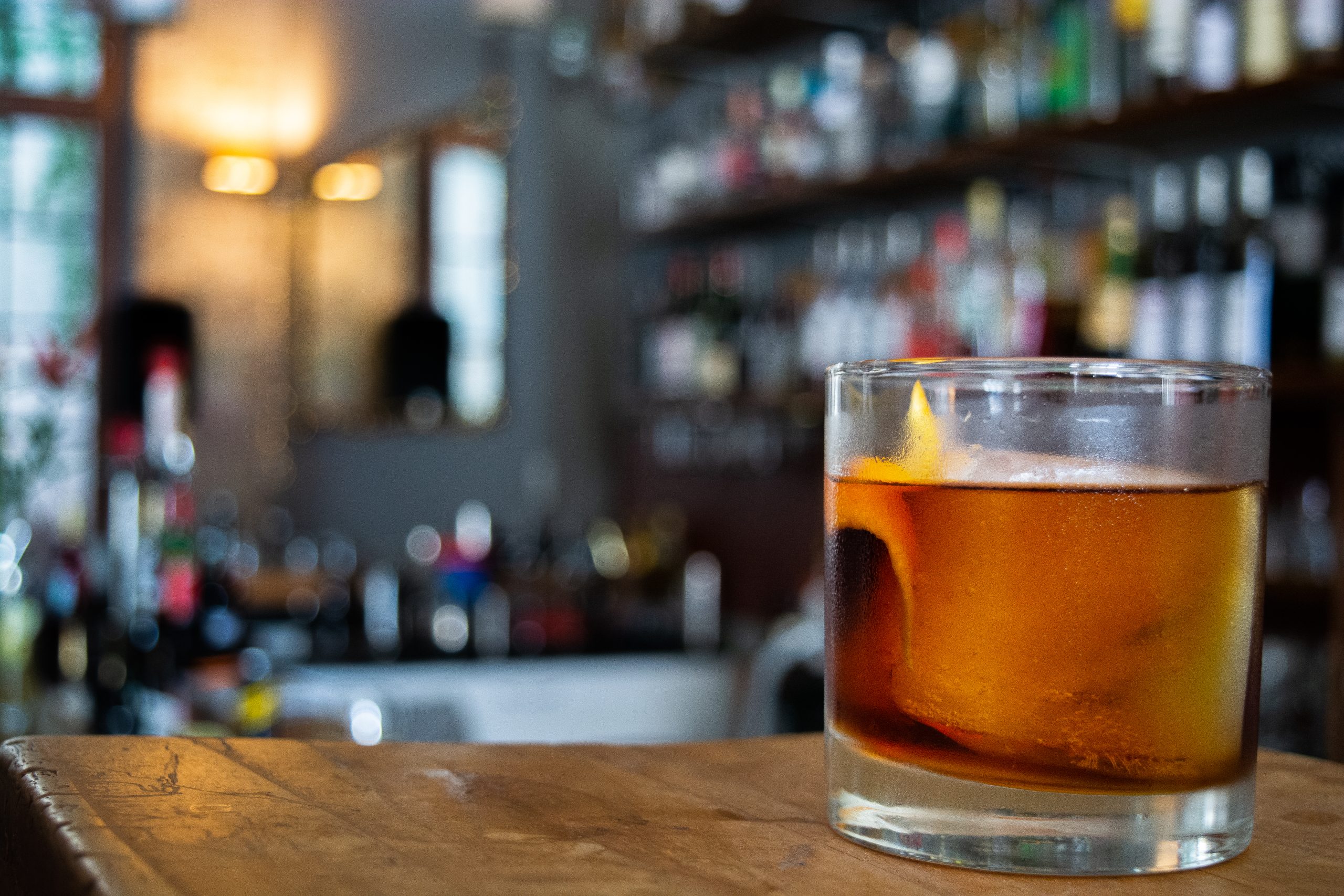a whiskey cocktail on bar bench with bottles blurred in background