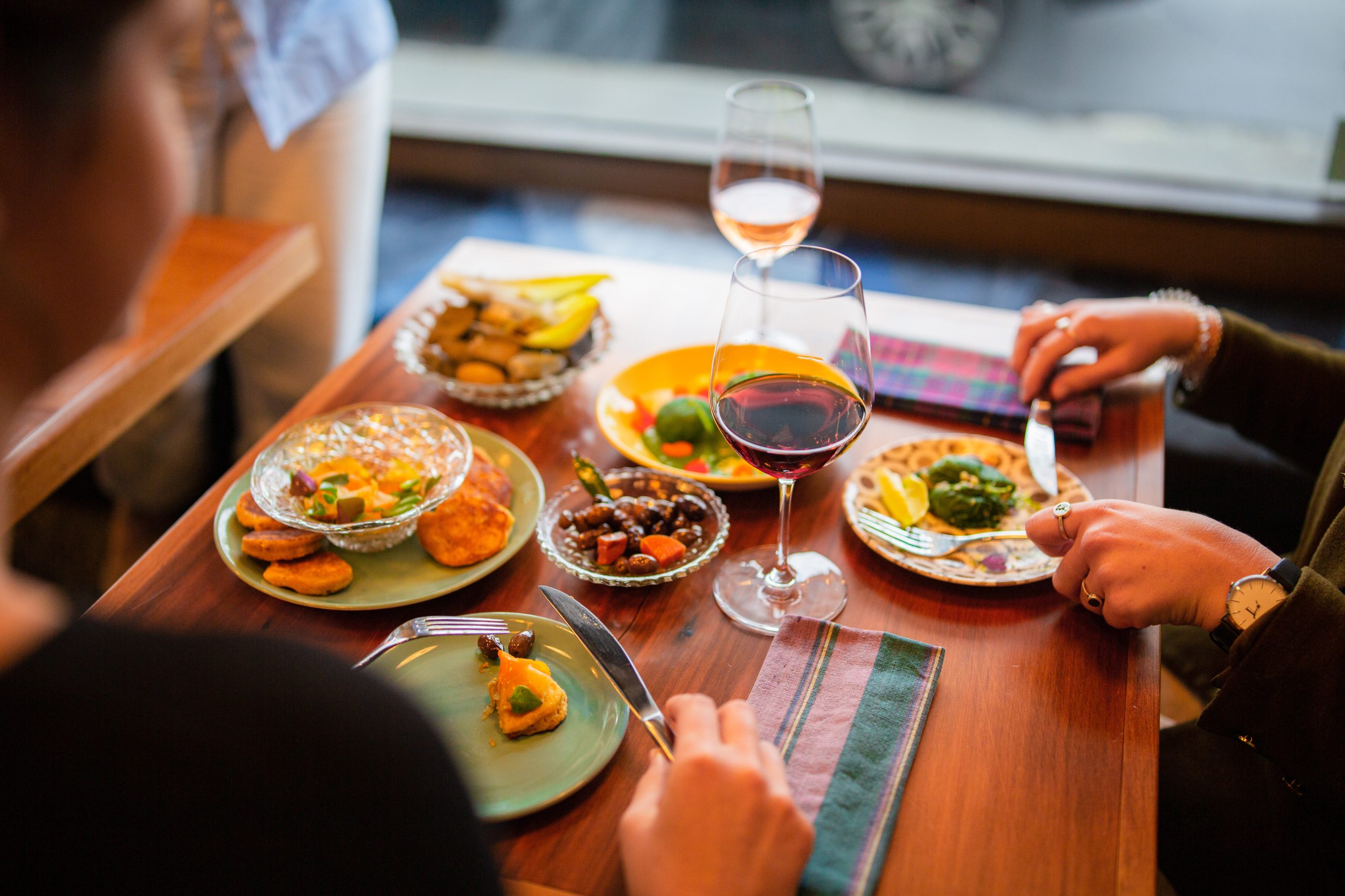 close up of 5 resturant dishes on table with hands and cutlery in view and wine glass by window